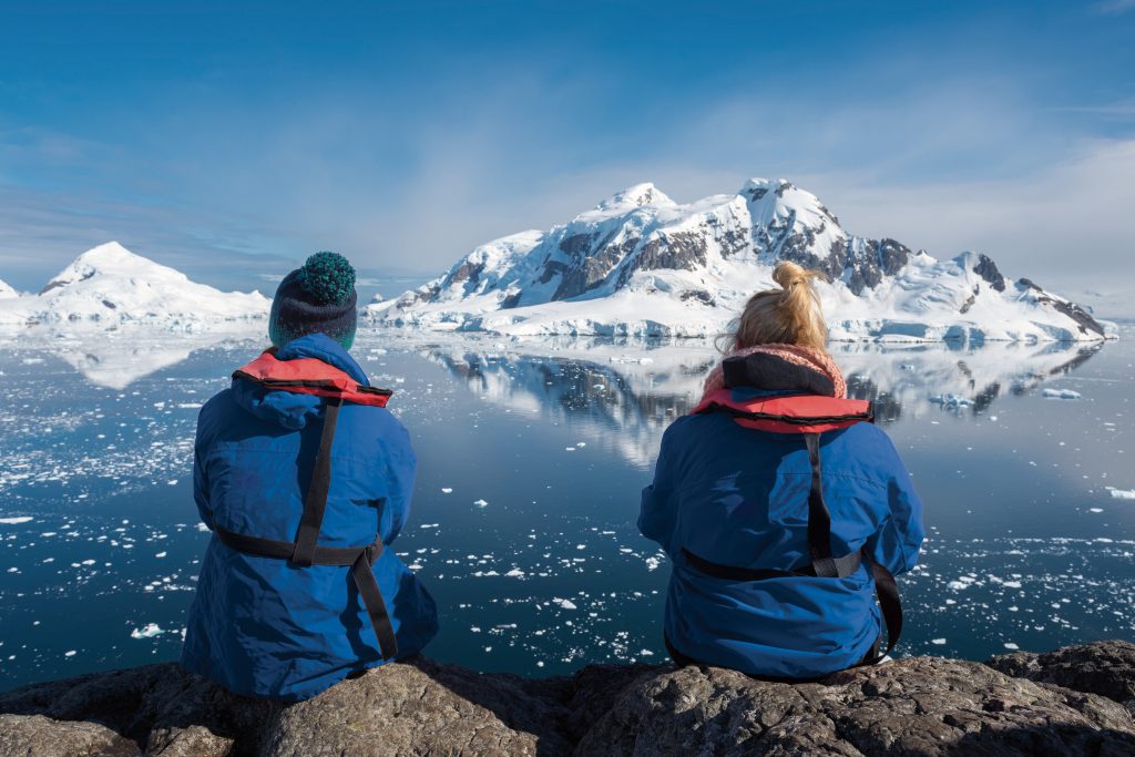 Two explorers sit side by side overlooking icy ocean and mountains